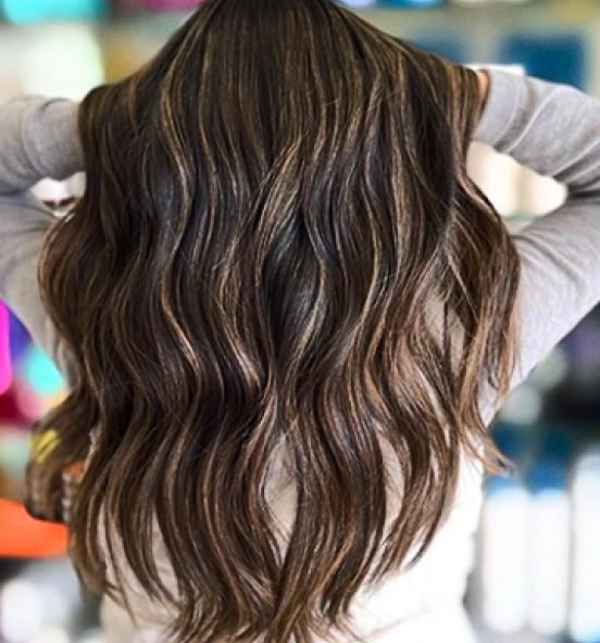 Person with long, wavy brown hair with highlights, viewed from the back with hands behind their head. Link goes to Hair Cuttery's Instagram page.